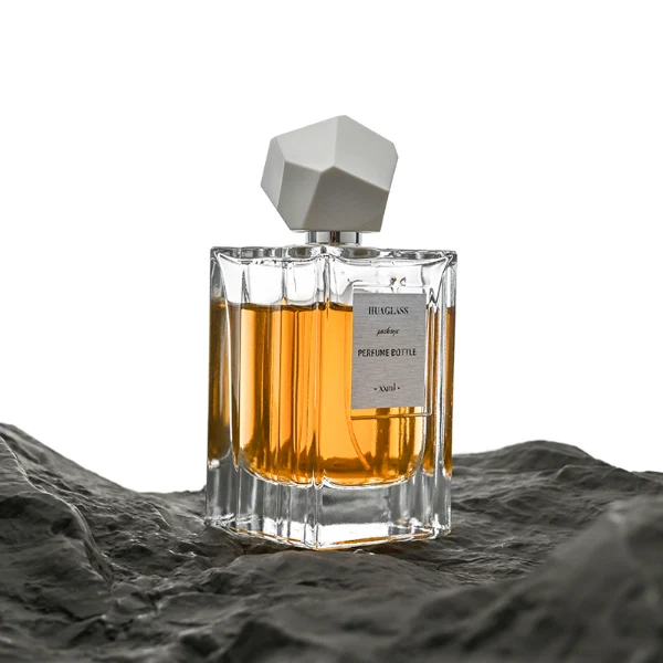 HUA0025 perfume bottle, heavy-base square cuboid glass bottle with fluted side details and clear finish, featuring a square paper label and an eye-catching irregular multi-faceted rock-shaped plastic cap with a high-gloss shiny finish and sharp geometric angles, elegant modern fragrance packaging design available in 50ml and 100ml