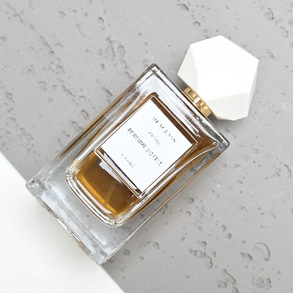 HUA0025 perfume bottle, heavy-base square cuboid glass bottle with fluted side details and clear finish, featuring a square paper label and an eye-catching irregular multi-faceted rock-shaped plastic cap with a high-gloss shiny finish and sharp geometric angles, elegant modern fragrance packaging design available in 50ml and 100ml