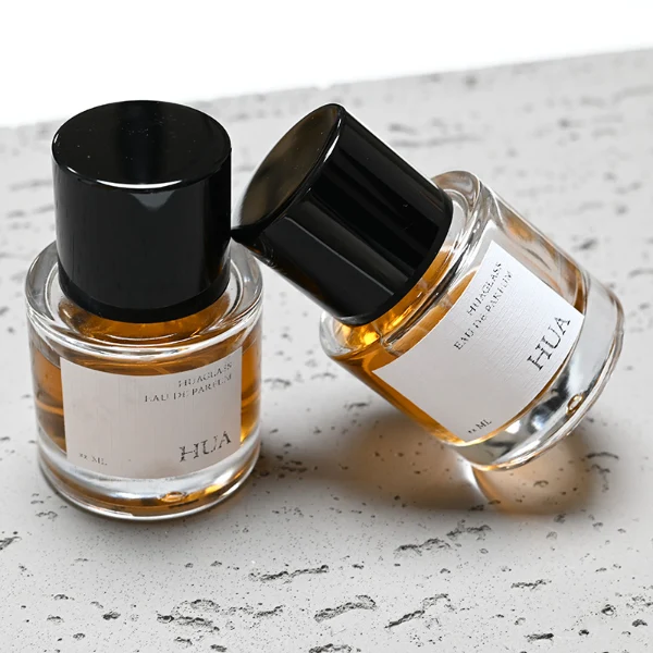 HUA0018 perfume bottle, heavy-base cylindrical round glass bottle with clear finish, featuring a rectangular paper label and a tall cylindrical plastic cap with a high-gloss black finish, elegant modern fragrance packaging design available in 30ml, 50ml, and 100ml
