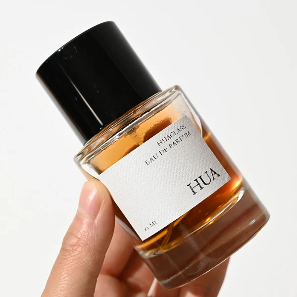 HUA0047 perfume bottle, heavy-base cylindrical round glass bottle with clear finish, featuring a square paper label and a wide cylindrical solid black plastic cap with a smooth high-gloss shiny finish, elegant modern fragrance packaging design available in 30ml, 50ml, and 100ml