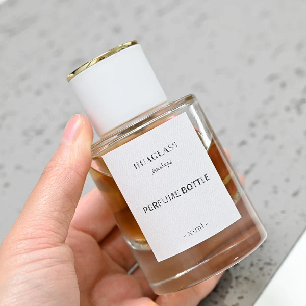 HUA0037 perfume bottle, heavy-base cylindrical round glass bottle with clear finish, featuring a square paper label, a metallic gold collar neck accent, and a short cylindrical plastic cap with a smooth high-gloss shiny finish, elegant modern fragrance packaging design available in 30ml, 50ml, and 100ml
