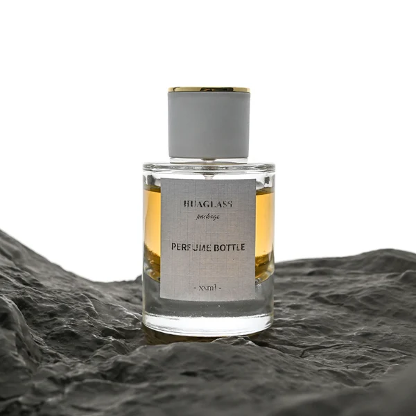 HUA0037 perfume bottle, heavy-base cylindrical round glass bottle with clear finish, featuring a square paper label, a metallic gold collar neck accent, and a short cylindrical plastic cap with a smooth high-gloss shiny finish, elegant modern fragrance packaging design available in 30ml, 50ml, and 100ml