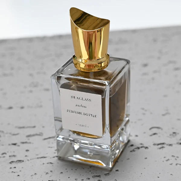 HUA0058 perfume bottle, heavy-base rectangular cuboid glass bottle with clear finish, featuring a paper label, a matching neck, and a tall sculptural cylindrical plastic cap with a sharply angled sloping top in a smooth high-gloss shiny gold finish, elegant modern fragrance packaging design available in 30ml, 50ml, and 100ml