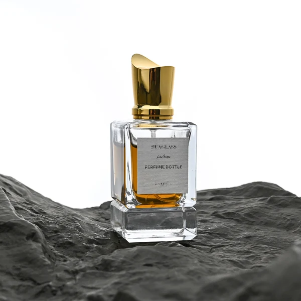 HUA0058 perfume bottle, heavy-base rectangular cuboid glass bottle with clear finish, featuring a paper label, a matching neck, and a tall sculptural cylindrical plastic cap with a sharply angled sloping top in a smooth high-gloss shiny gold finish, elegant modern fragrance packaging design available in 30ml, 50ml, and 100ml