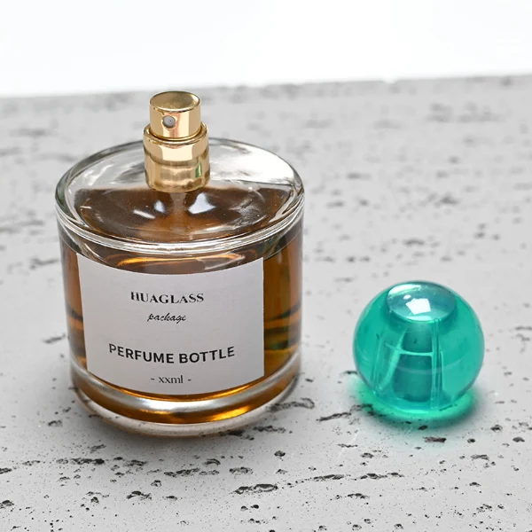 HUA0028 perfume bottle, transparent colorless rounded cylindrical glass body with thick base and smooth curved shoulder, 100 ml liquid, spherical transparent plastic cap with visible inner spray structure in green or clear color, brand label attached on bottle surface, modern elegant luxury design, ideal for gifting and daily use.