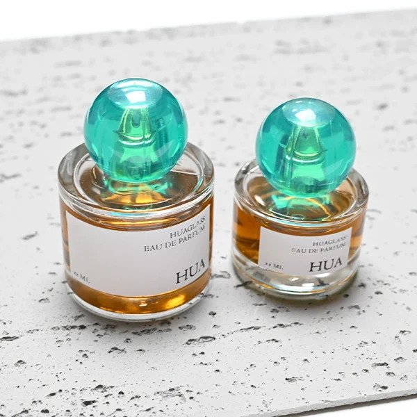 HUA0028 perfume bottle, transparent colorless rounded cylindrical glass body with thick base and smooth curved shoulder, 100 ml liquid, spherical transparent plastic cap with visible inner spray structure in green or clear color, brand label attached on bottle surface, modern elegant luxury design, ideal for gifting and daily use.