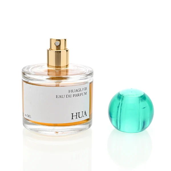 HUA0028 perfume bottle, transparent colorless rounded cylindrical glass body with thick base and smooth curved shoulder, 100 ml liquid, spherical transparent plastic cap with visible inner spray structure in green or clear color, brand label attached on bottle surface, modern elegant luxury design, ideal for gifting and daily use.