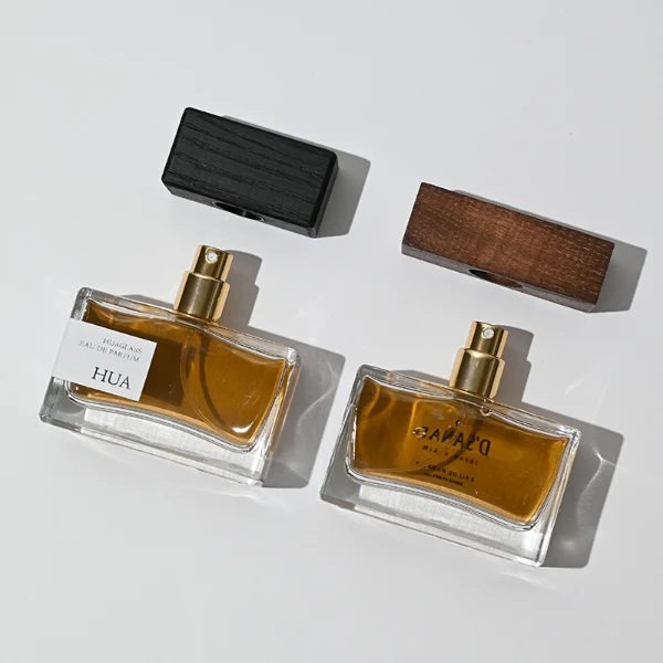 HUA0036 perfume bottle, heavy-base wide rectangular cuboid glass bottle with clear finish, featuring a side-set paper label, a metallic gold collar neck accent, and a prominent wide rectangular block-shaped wood cap with prominent natural wood grain lines, elegant modern fragrance packaging design available in 30ml, 50ml, and 100ml