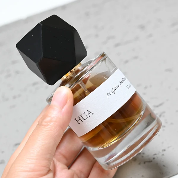 HUA0053 perfume bottle, heavy-base cylindrical round glass bottle with clear finish, featuring a wide wrapped paper label, a gold collar neck accent, and a unique polyhedral faceted diamond-shaped wood cap with subtle grain textures, elegant modern fragrance packaging design available in 30ml, 50ml, and 100ml