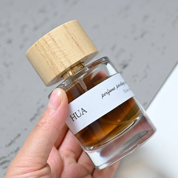 HUA0035 perfume bottle, heavy-base cylindrical round glass bottle with clear finish, featuring a horizontal paper label, a polished gold neck, and a wide cylindrical timber cap with a natural smooth grain finish, elegant modern fragrance packaging design available in 30ml, 50ml, and 100ml