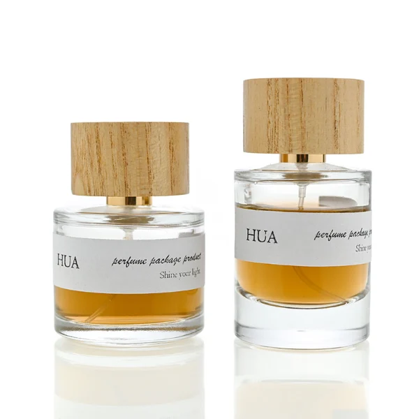 HUA0035 perfume bottle, heavy-base cylindrical round glass bottle with clear finish, featuring a horizontal paper label, a polished gold neck, and a wide cylindrical timber cap with a natural smooth grain finish, elegant modern fragrance packaging design available in 30ml, 50ml, and 100ml