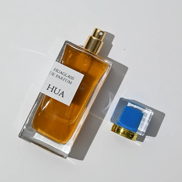 HUA0043 perfume bottle, heavy-base rectangular cuboid glass bottle with clear finish, featuring a side-aligned square paper label, a gold collar neck accent, and a unique double-layered square plastic cap with a transparent outer block and a colorful high-gloss shiny inner core, elegant modern fragrance packaging design available in 30ml, 50ml, and 100ml
