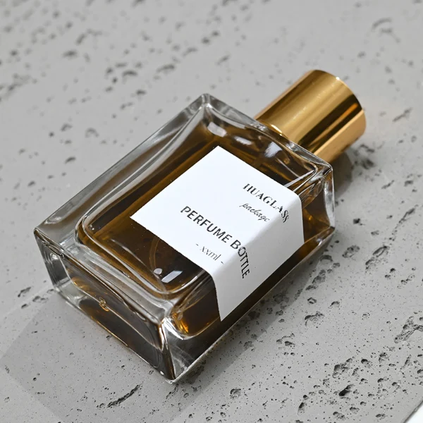 HUA0049 perfume bottle, heavy-base rectangular cuboid glass bottle with clear finish, featuring a corner-wrapped square paper label, a coordinating collar neck accent, and a tall cylindrical aluminum cap with a smooth polished metallic finish, elegant modern fragrance packaging design available in 30ml, 50ml, and 100ml