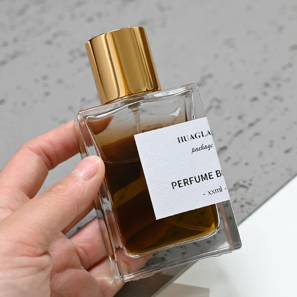 HUA0049 perfume bottle, heavy-base rectangular cuboid glass bottle with clear finish, featuring a corner-wrapped square paper label, a coordinating collar neck accent, and a tall cylindrical aluminum cap with a smooth polished metallic finish, elegant modern fragrance packaging design available in 30ml, 50ml, and 100ml