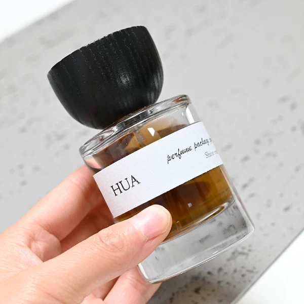 HUA0032 perfume bottle, heavy-base cylindrical round glass bottle with clear finish, featuring a horizontal paper label, a matching neck, and an inverted conical timber cap with a natural smooth grain finish, elegant modern fragrance packaging design available in 30ml, 50ml, and 100ml