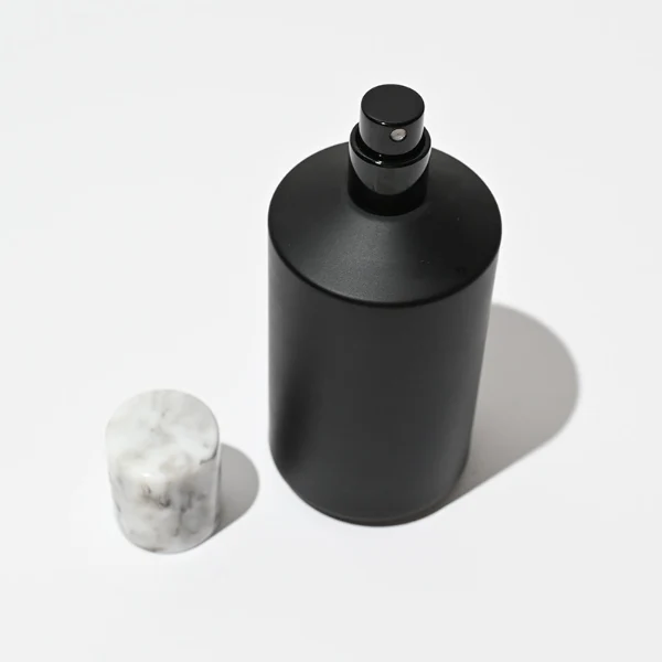 HUA0038 perfume bottle, heavy-base cylindrical round glass bottle with clear or solid high-gloss opaque black finish and unique chamfered sloping shoulders, featuring a tall cylindrical resin cap with an eye-catching marbled texture, elegant modern fragrance packaging design available in 30ml, 50ml, and 100ml