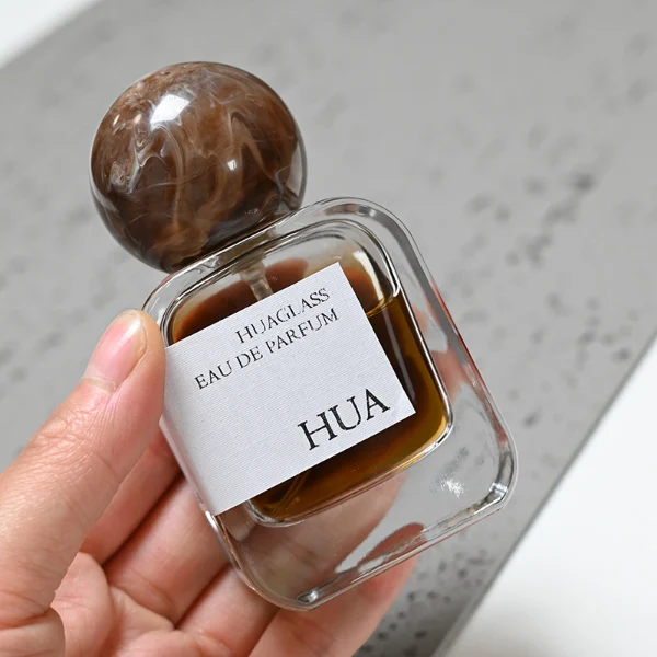 HUA0042 perfume bottle, clear flattened oval flask glass bottle featuring a prominent spherical ball-shaped resin cap with an eye-catching marbled texture and organic fluid swirls, modern playful fragrance packaging design available in 30ml, 50ml, and 100ml