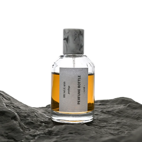 HUA0031 perfume bottle, heavy-base cylindrical glass bottle with a faceted shoulder and clear finish, featuring a square paper label, a matching neck, and a tall cylindrical resin cap with a colorful marbled finish, elegant modern fragrance packaging design available in 30ml, 50ml, and 100ml