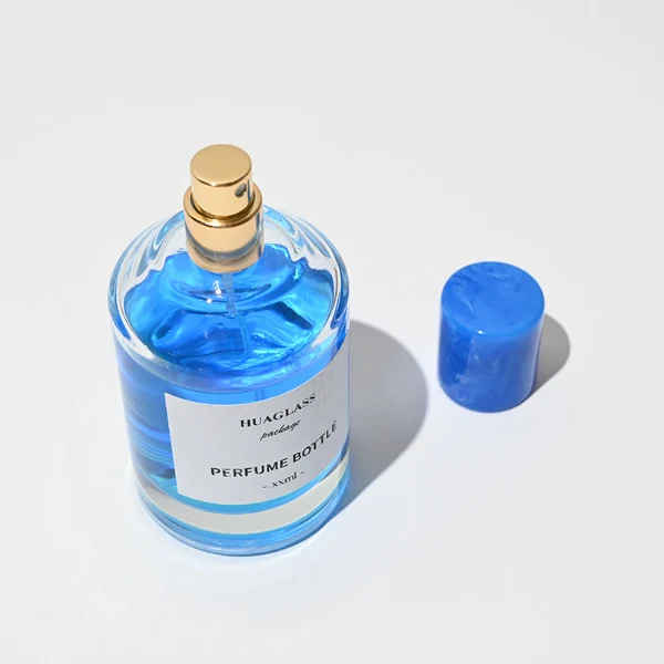 HUA0031 perfume bottle, heavy-base cylindrical glass bottle with a faceted shoulder and clear finish, featuring a square paper label, a matching neck, and a tall cylindrical resin cap with a colorful marbled finish, elegant modern fragrance packaging design available in 30ml, 50ml, and 100ml