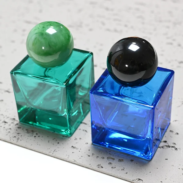 HUA0009 perfume bottle, luxury heavy-base square cuboid glass bottle with translucent deep multi-color finish, featuring a prominent oversized spherical solid colored resin cap showcasing opaque organic swirling veining and high-gloss textures, contemporary elegant fragrance packaging design available in 30ml, 50ml, and 100ml