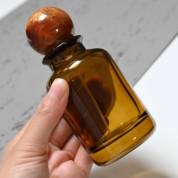 HUA0044 perfume bottle, cylindrical round glass bottle in high-transparency amber or solid high-gloss opaque black finish, featuring a prominent spherical ball-shaped resin cap with an eye-catching marbled texture and organic fluid swirls, elegant modern fragrance packaging design available in 30ml, 50ml, and 100ml