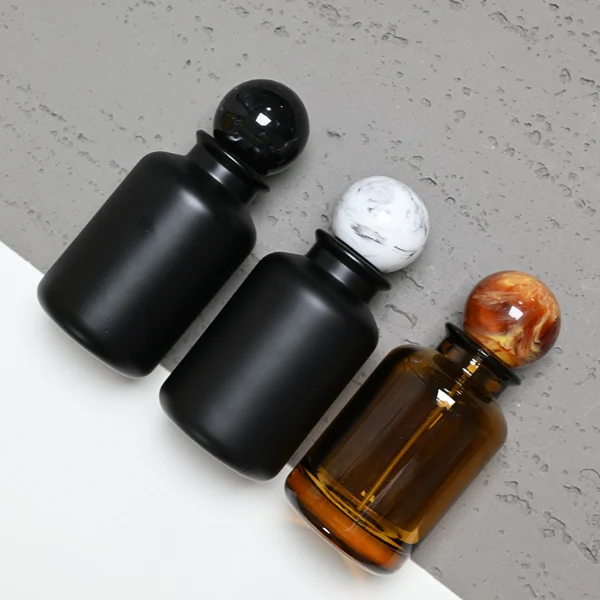 HUA0044 perfume bottle, cylindrical round glass bottle in high-transparency amber or solid high-gloss opaque black finish, featuring a prominent spherical ball-shaped resin cap with an eye-catching marbled texture and organic fluid swirls, elegant modern fragrance packaging design available in 30ml, 50ml, and 100ml