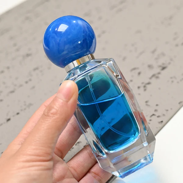 HUA0034 perfume bottle, heavy-base cylindrical glass bottle with a faceted shoulder and clear finish, featuring a square paper label, a polished gold or silver neck, and a tall cylindrical resin cap with a colorful marbled finish, elegant modern fragrance packaging design available in 50ml