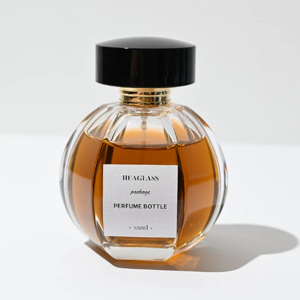 HUA0012 perfume bottle, heavy-base ribbed spherical round glass bottle with clear finish, featuring a square paper label, a matching neck, and a wide cylindrical plastic cap with a smooth high-gloss shiny black finish, elegant modern fragrance packaging design available in 100ml