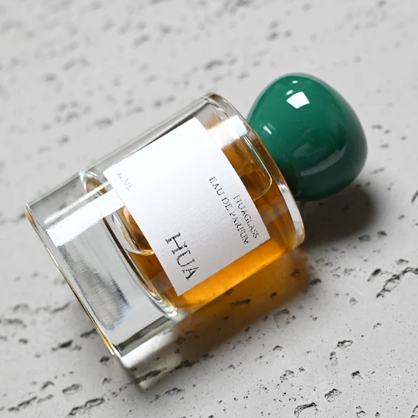 HUA0026 perfume bottle, heavy-base cylindrical round glass bottle with clear finish, featuring a square paper label and an organic smooth pebble-shaped plastic cap with a high-gloss shiny finish, elegant modern fragrance packaging design available in 30ml, 50ml, and 100ml