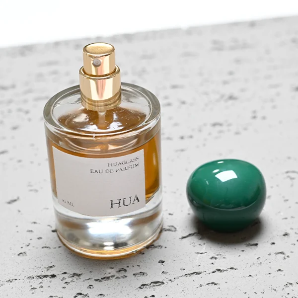 HUA0026 perfume bottle, heavy-base cylindrical round glass bottle with clear finish, featuring a square paper label and an organic smooth pebble-shaped plastic cap with a high-gloss shiny finish, elegant modern fragrance packaging design available in 30ml, 50ml, and 100ml