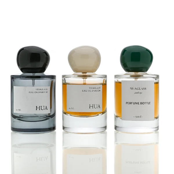 HUA0026 perfume bottle, heavy-base cylindrical round glass bottle with clear finish, featuring a square paper label and an organic smooth pebble-shaped plastic cap with a high-gloss shiny finish, elegant modern fragrance packaging design available in 30ml, 50ml, and 100ml