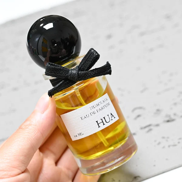 HUA0062 perfume bottle, heavy-base cylindrical round glass bottle with clear finish, featuring a square paper label, a matching neck with ribbon bow decorations, and a playful spherical ball-shaped plastic cap with a smooth high-gloss shiny finish, elegant modern fragrance packaging design available in 30ml, 50ml, and 100ml