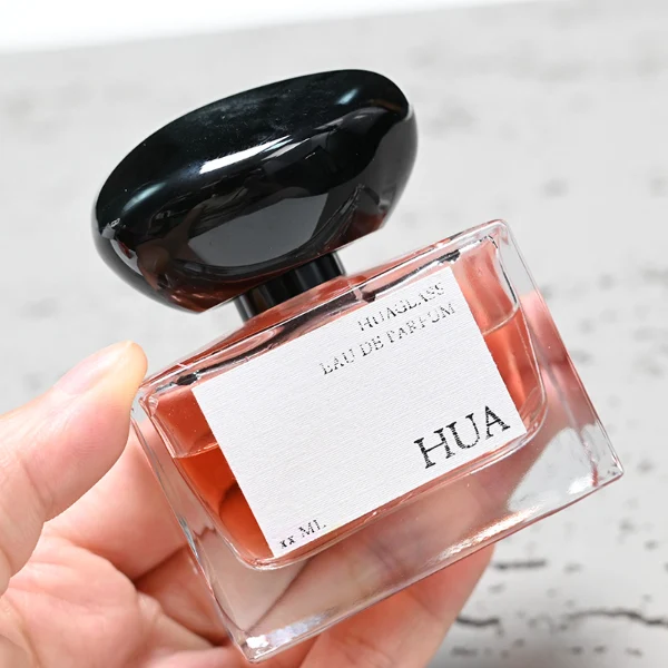 HUA0096 perfume bottle, rectangular glass body, glossy black pebble-shaped plastic cap with yellow accents, modern minimalist design, available in 30 ml, 50 ml, and 100 ml.