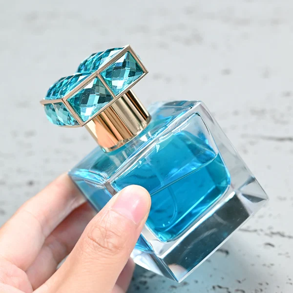 HUA0100 perfume bottle, square glass body with liquid, rectangular geometric zinc alloy cap with colorful crystal detailing on the top, modern luxurious design, available in 30 ml, 50 ml, and 100 ml.