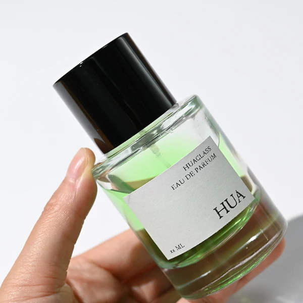 HUA0108 perfume bottle, cylindrical glass body with liquid and outer label, glossy black plastic cylindrical cap with silver metallic neck, modern elegant design, available in 30 ml, 50 ml, and 100 ml.