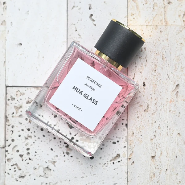 HUA0111 perfume bottle, square glass body with liquid and outer label, cylindrical plastic cap with gold metallic neck, modern minimalist design, available in 30 ml, 50 ml, and 100 ml.