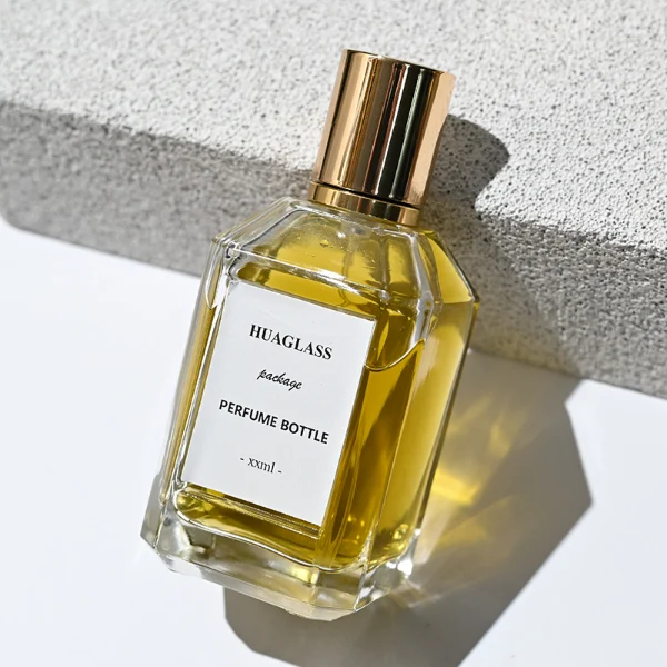 HUA0135 perfume bottle, rectangular clear glass body with 30 ml, 50 ml, or 100 ml liquid, cylindrical plastic cap with gold metallic neck, outer label with printed logo, modern elegant design, ideal for gifting and daily use.