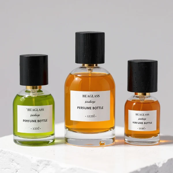 HUA0162 perfume bottle, cylindrical transparent glass body with 30 ml, 50 ml, or 100 ml liquid, tall cylindrical black wooden cap with gold metallic neck, outer label with printed logo, modern elegant design, ideal for gifting and daily use.