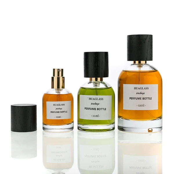 HUA0162 perfume bottle, cylindrical transparent glass body with 30 ml, 50 ml, or 100 ml liquid, tall cylindrical black wooden cap with gold metallic neck, outer label with printed logo, modern elegant design, ideal for gifting and daily use.