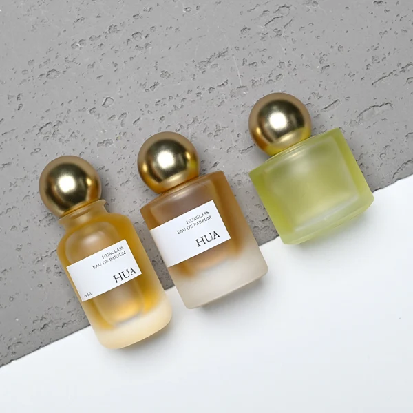 HUA0164 perfume bottle, cylindrical frosted glass body with 30 ml, 50 ml, or 100 ml liquid, spherical gold-colored plastic cap, outer label with printed logo, modern elegant design, ideal for gifting and daily use.