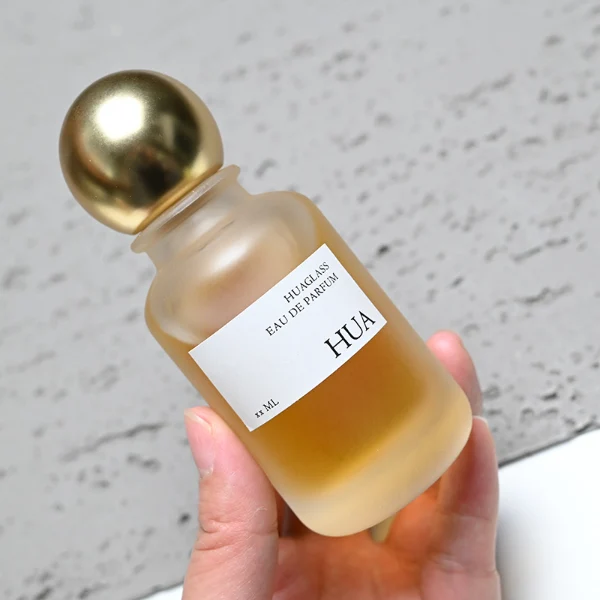 HUA0164 perfume bottle, cylindrical frosted glass body with 30 ml, 50 ml, or 100 ml liquid, spherical gold-colored plastic cap, outer label with printed logo, modern elegant design, ideal for gifting and daily use.
