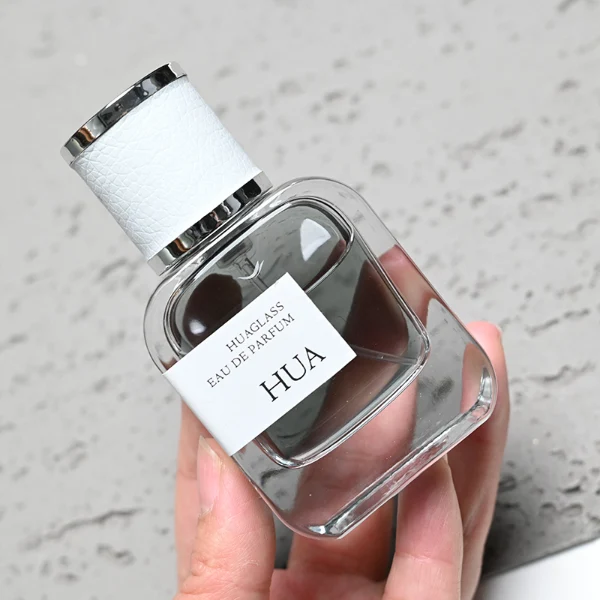 HUA0165 perfume bottle, rectangular transparent glass body with 30 ml, 50 ml, or 100 ml liquid, cylindrical plastic cap wrapped in leather with gold metallic neck, outer label with printed logo, modern elegant design, ideal for gifting and daily use.