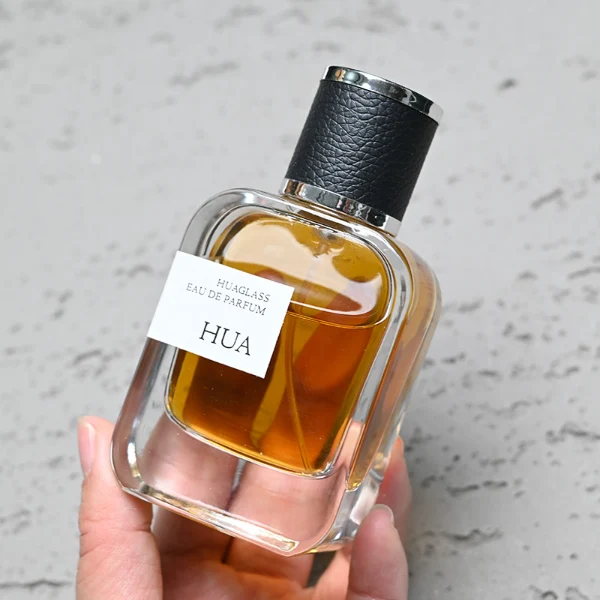 HUA0165 perfume bottle, rectangular transparent glass body with 30 ml, 50 ml, or 100 ml liquid, cylindrical plastic cap wrapped in leather with gold metallic neck, outer label with printed logo, modern elegant design, ideal for gifting and daily use.