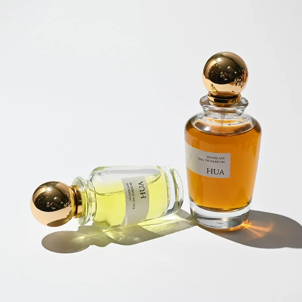 HUA0167 perfume bottle, kettle-shaped cylindrical transparent glass body with 50 ml or 100 ml liquid, spherical glossy plastic cap with gold neck, outer label with printed logo, modern elegant design, ideal for gifting and daily use.