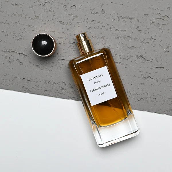 HUA0170 perfume bottle, rectangular transparent glass body with 50 ml or 80 ml liquid, cylindrical plastic cap with engraved decorative pattern and gold neck ring, outer label with printed logo, modern luxurious design, ideal for gifting and daily use.
