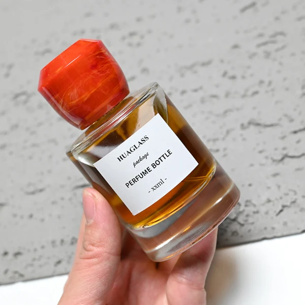 HUA0174 perfume bottle, cylindrical transparent glass body with 30 ml, 50 ml, or 100 ml liquid, faceted geometric plastic cap with marble-effect appearance, printed logo label, modern artistic luxury design, ideal for gifting and daily use.