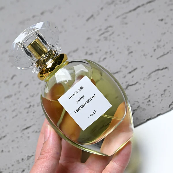 HUA0183 perfume bottle, oval transparent glass body with 30 ml, 50 ml, 75 ml, or 100 ml liquid, transparent disc-shaped plastic cap with layered decorative neck detail, printed logo label, modern elegant luxury design, ideal for gifting and daily use.
