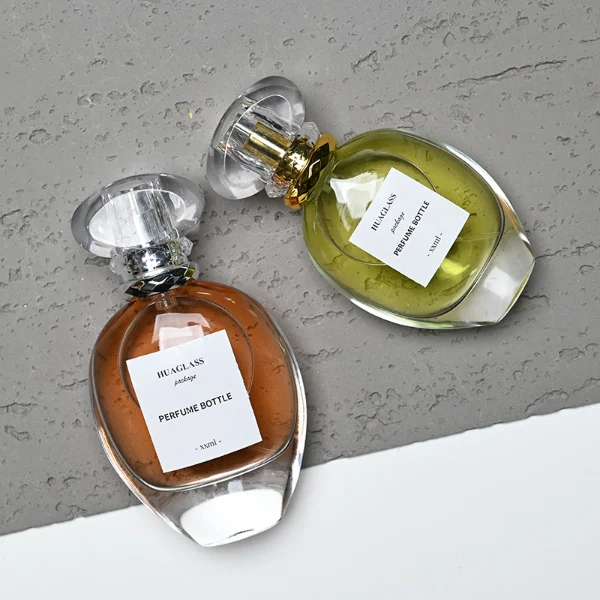 HUA0183 perfume bottle, oval transparent glass body with 30 ml, 50 ml, 75 ml, or 100 ml liquid, transparent disc-shaped plastic cap with layered decorative neck detail, printed logo label, modern elegant luxury design, ideal for gifting and daily use.