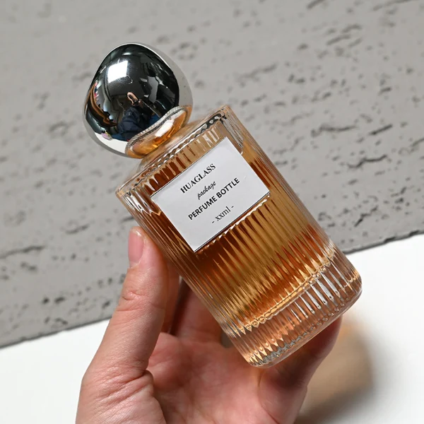 HUA0184 perfume bottle, cylindrical ribbed transparent glass body with 50 ml or 100 ml liquid, irregular glossy spherical plastic cap with customizable colors, printed logo label, modern artistic luxury design, ideal for gifting and daily use.