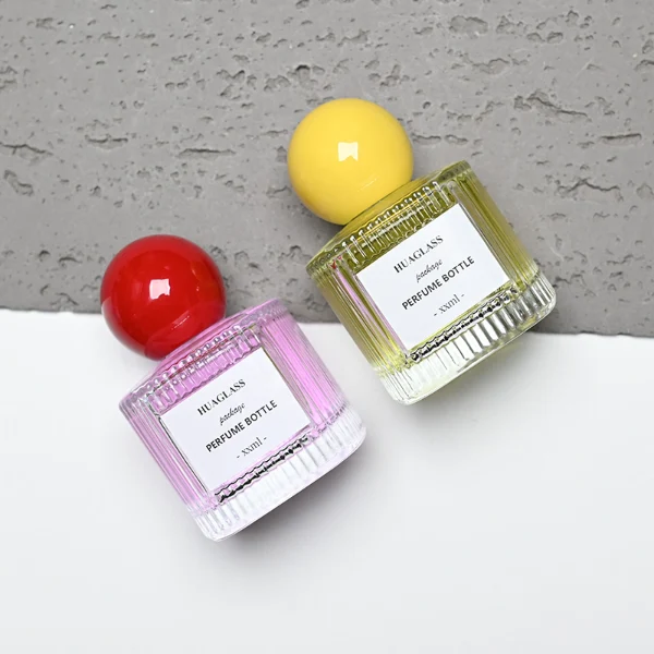 HUA0203 perfume bottle, cylindrical ribbed transparent glass body with thick base, 50 ml or 100 ml liquid, spherical glossy plastic cap in red or yellow, brand label applied on the surface, modern luxurious design, ideal for gifting and daily use.